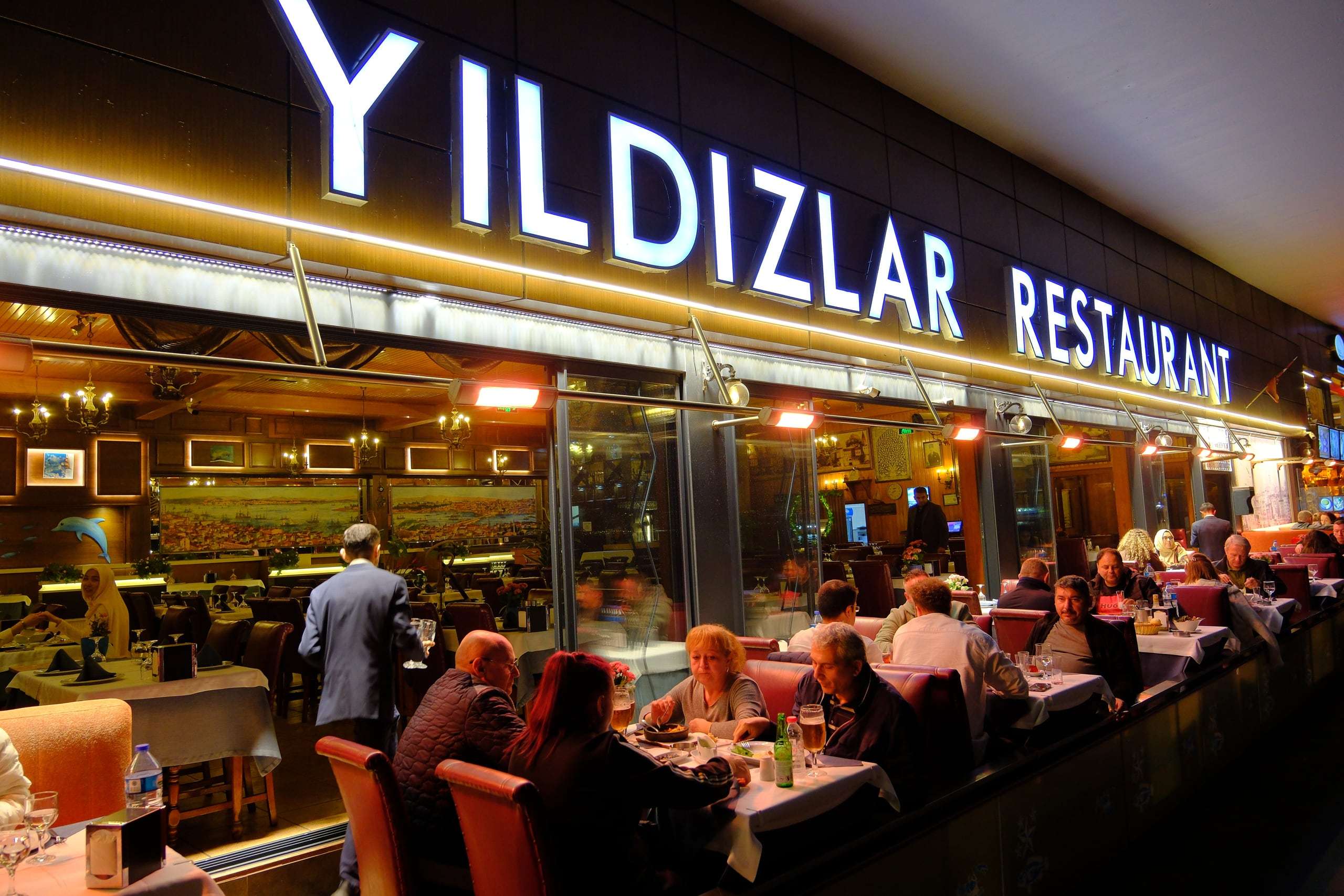 Lit restaurant sign under the Galata Bridge at night with full tables along the water